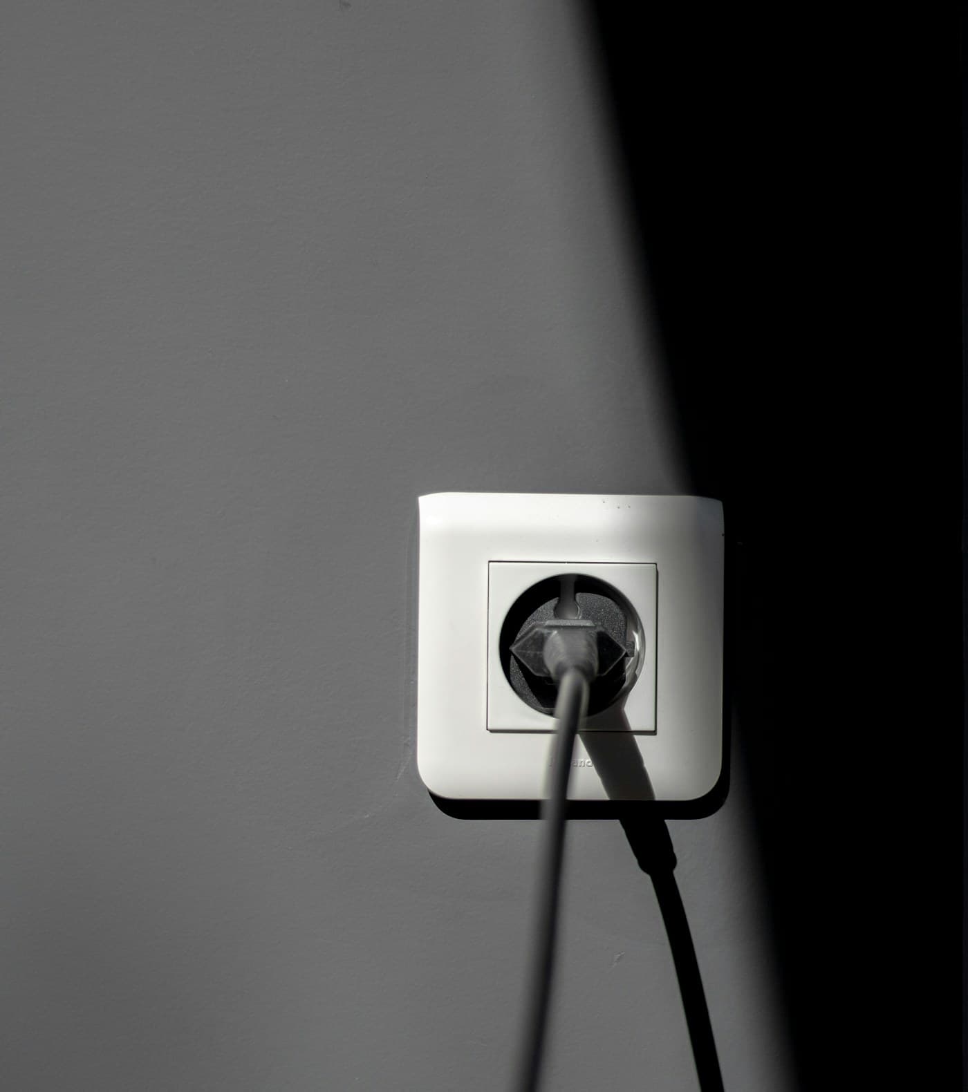 Une prise de courant européenne sur un mur gris, traversée par un trait de lumière, avec son câble branché qui retombe.